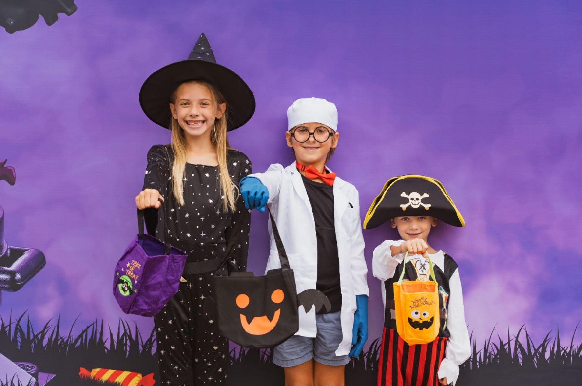 Three children in Halloween costumes—a witch, a scientist, and a pirate—stand in front of a purple background holding trick-or-treat bags.