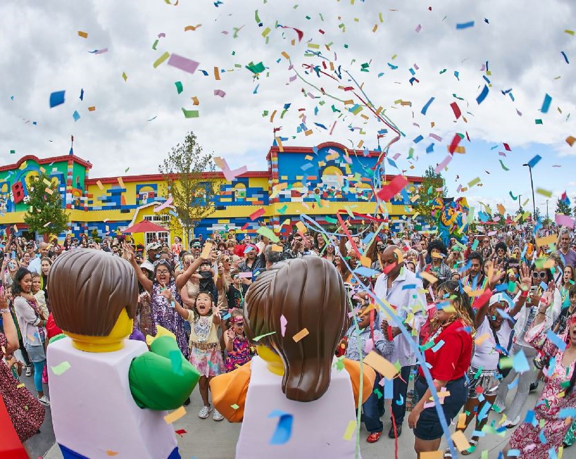 A crowd of people and children celebrate outdoors with confetti, joined by two large LEGO character mascots in front of a colorful building.