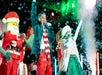 Performers in festive attire sing on stage with a snow effect, flanked by a person in a Santa costume and dancers in red and green striped outfits.