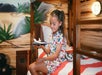 A young girl in printed pajamas sits on a bunk bed holding a plate with cake or dessert, in a room decorated with a colorful, tropical wall mural.
