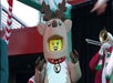 Person in a reindeer costume with a LEGO face poses, while a musician wearing a red and white outfit plays a trombone in the background.