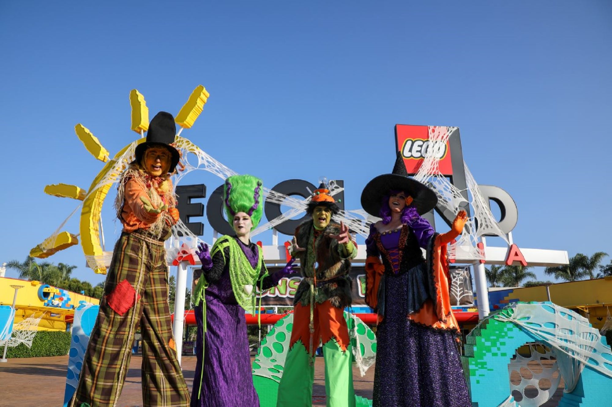 Four people in colorful witch costumes stand in front of a LEGOLAND sign decorated with Halloween-themed LEGO structures under a clear blue sky.