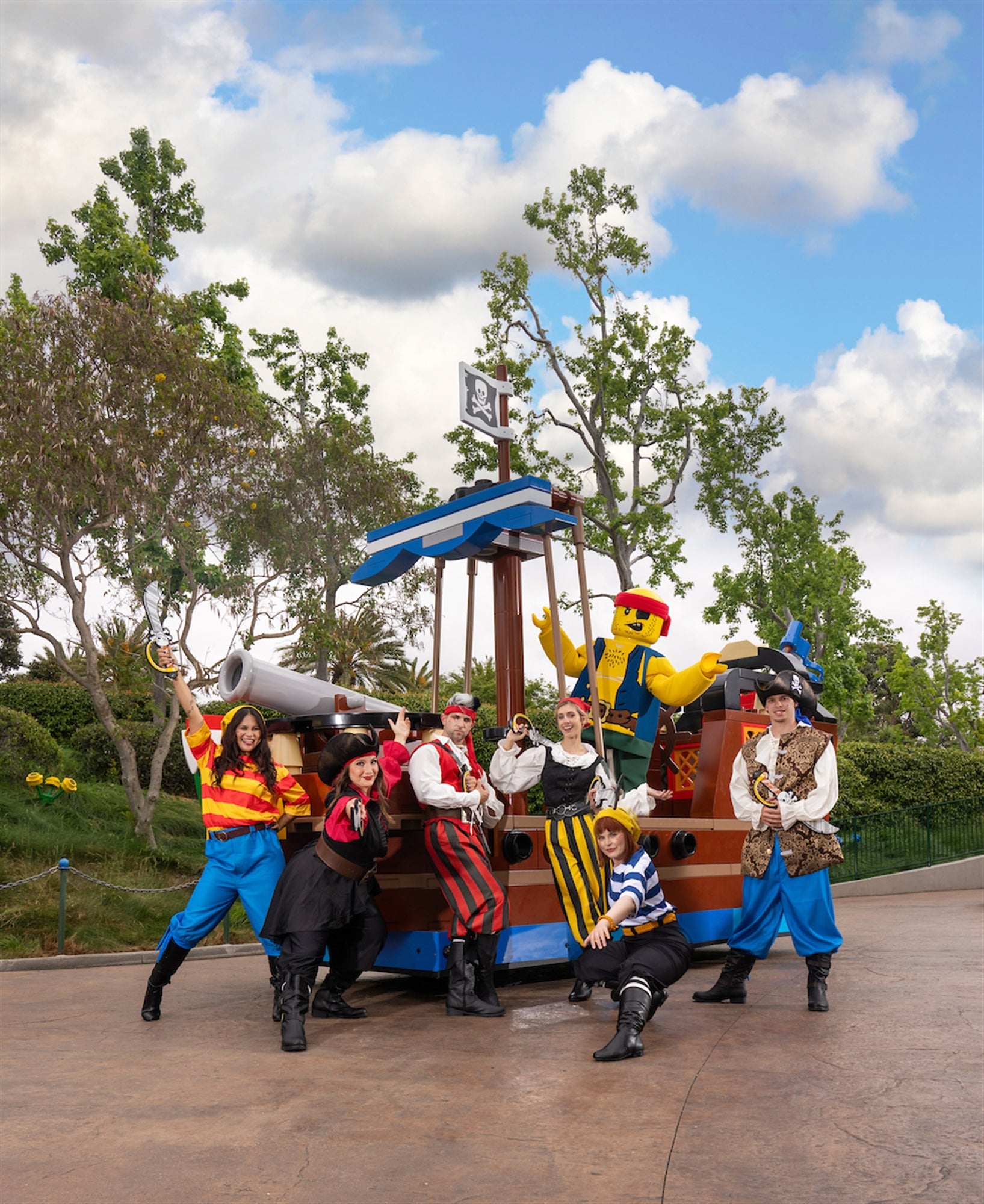 A group of people in colorful pirate costumes pose in front of a pirate-themed float with a large LEGO pirate figure outdoors.