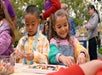 Two children sit at a table outdoors, focused on building with colorful plastic blocks, with adults and other children in the background.