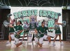 Cheerleaders in green uniforms pose with a person in a zombie LEGO costume onstage in front of a "Zombie Cheer Crew" sign.