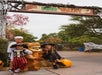Three children in Halloween costumes pose next to a dinosaur LEGO statue under a "Dino Valley" sign at an outdoor theme park decorated with pumpkins and spider webs.