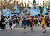Parade performers in colorful outfits wave blue flags while marching, as costumed staff and spectators watch along the street in an outdoor amusement park setting.