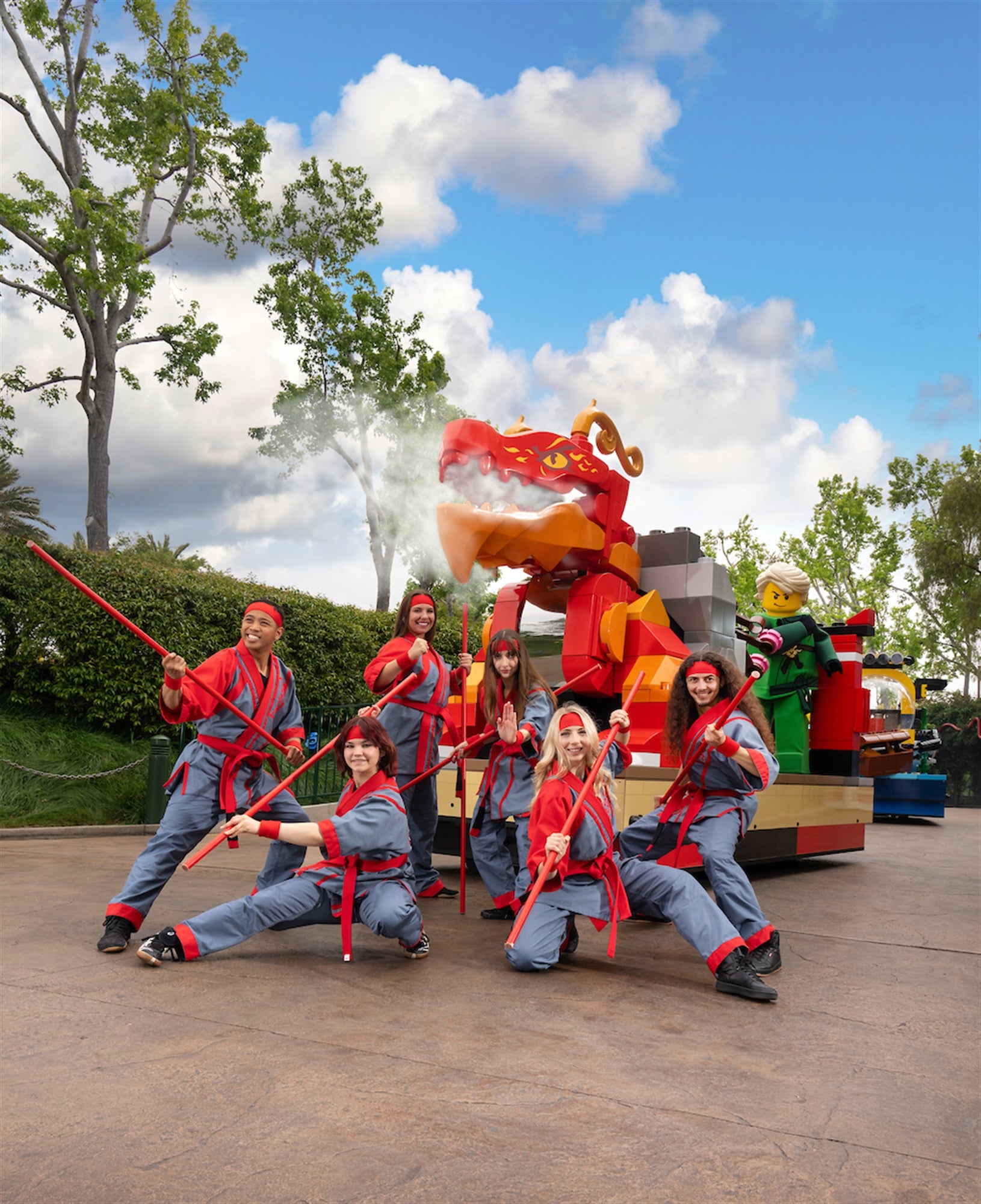 Six people in matching red and grey costumes pose with red staffs in front of a large LEGO dragon float outdoors on a sunny day.