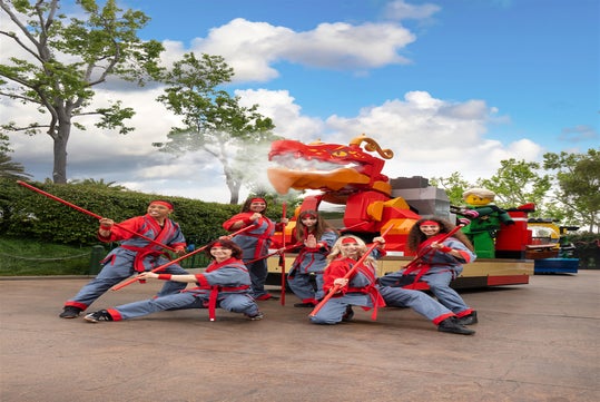 Six people in matching red and grey costumes pose with red staffs in front of a large LEGO dragon float outdoors on a sunny day.