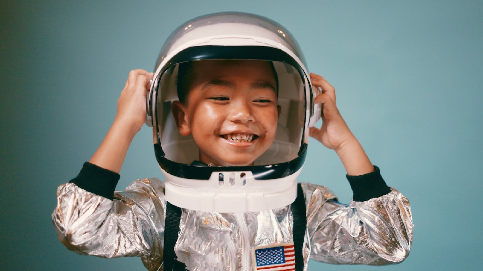 Smiling child wearing a silver astronaut suit and helmet with an American flag patch, standing against a blue background.