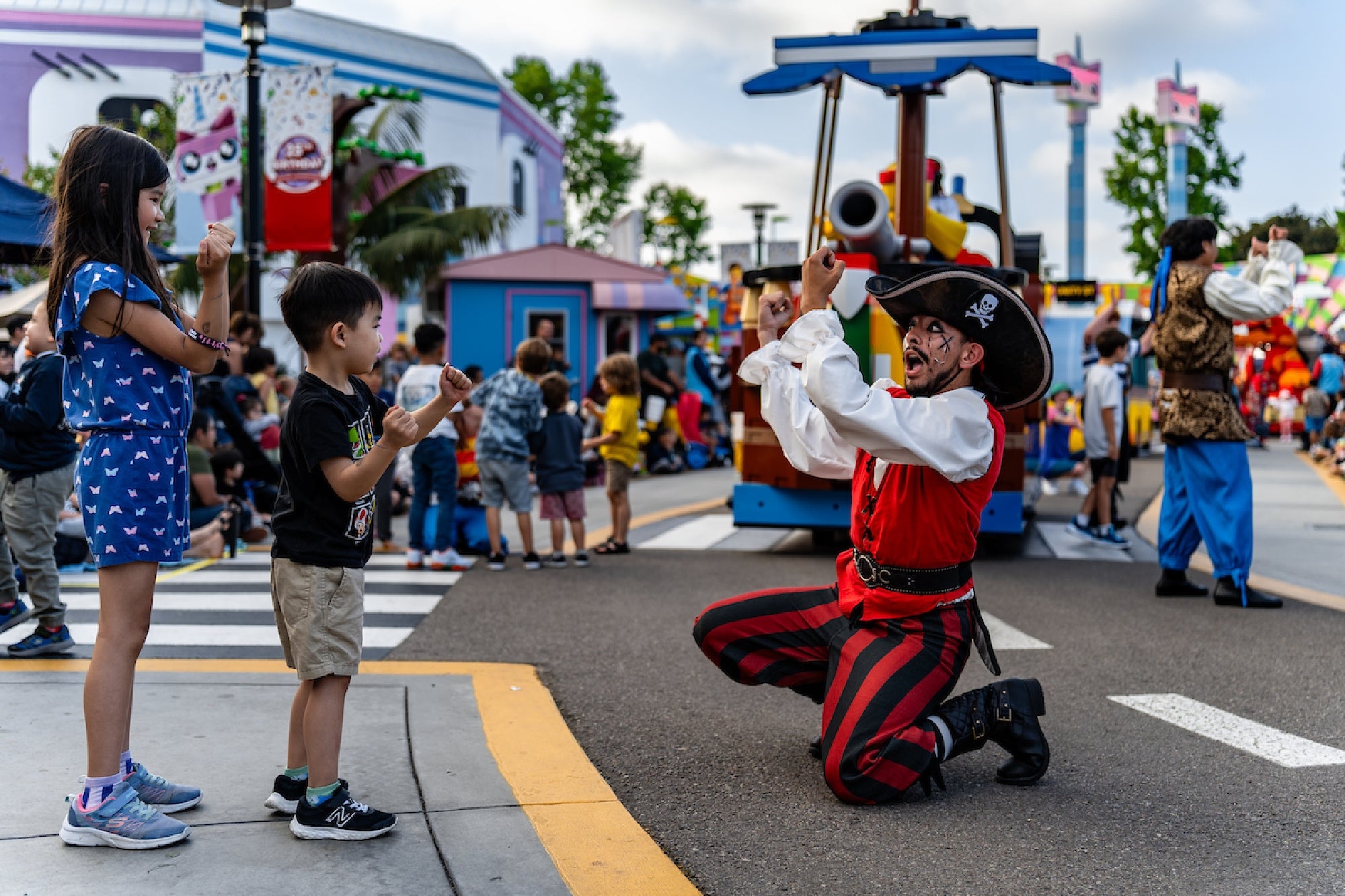A performer dressed as a pirate kneels and gestures to two children at an outdoor amusement park, with other people and colorful decorations in the background.