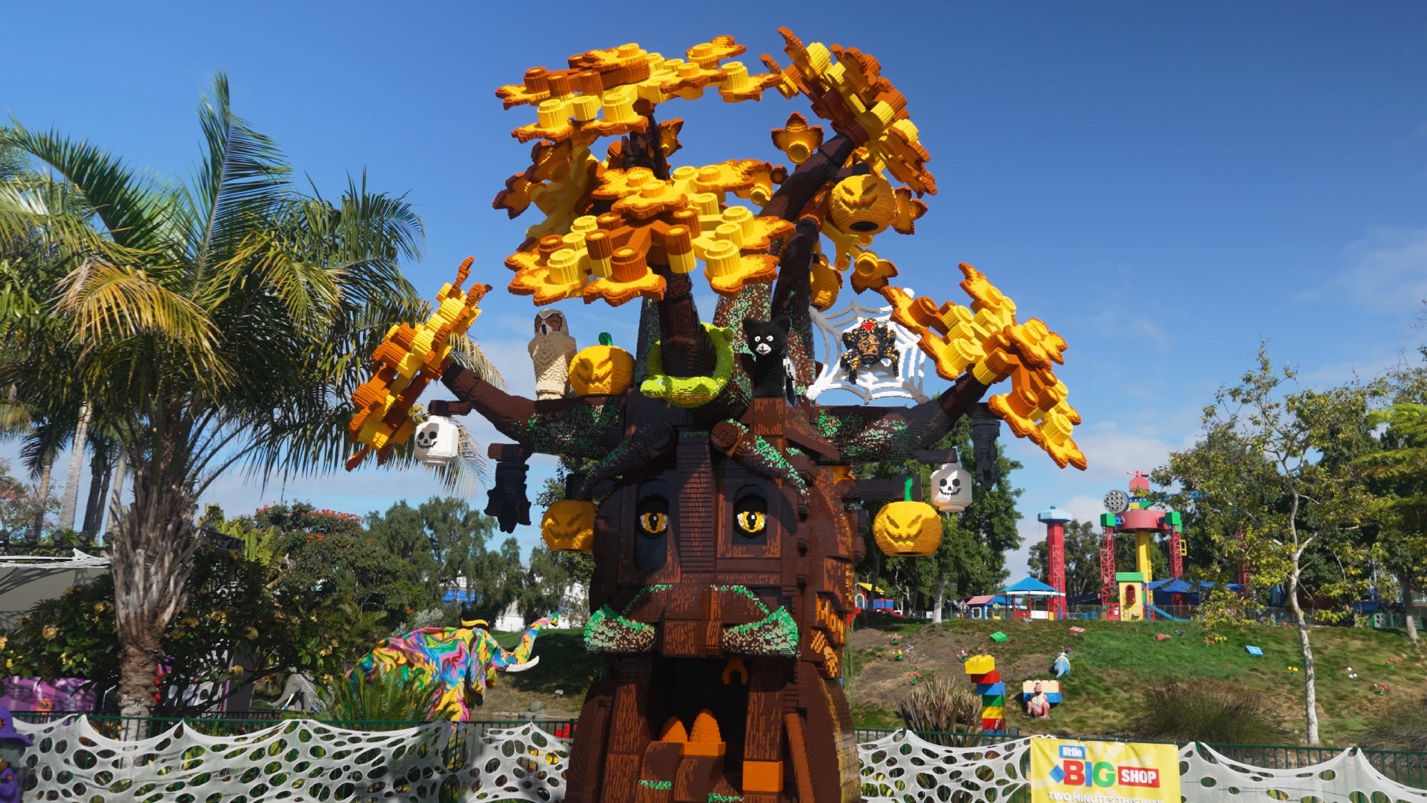 A large tree sculpture made of LEGO bricks features yellow and orange leaves, pumpkins, and animal faces. The scene is outdoors with trees, greenery, and a colorful building in the background.