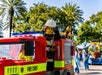 A person in a firefighter LEGO costume stands on a LEGO fire truck during an outdoor event with palm trees and people in the background.