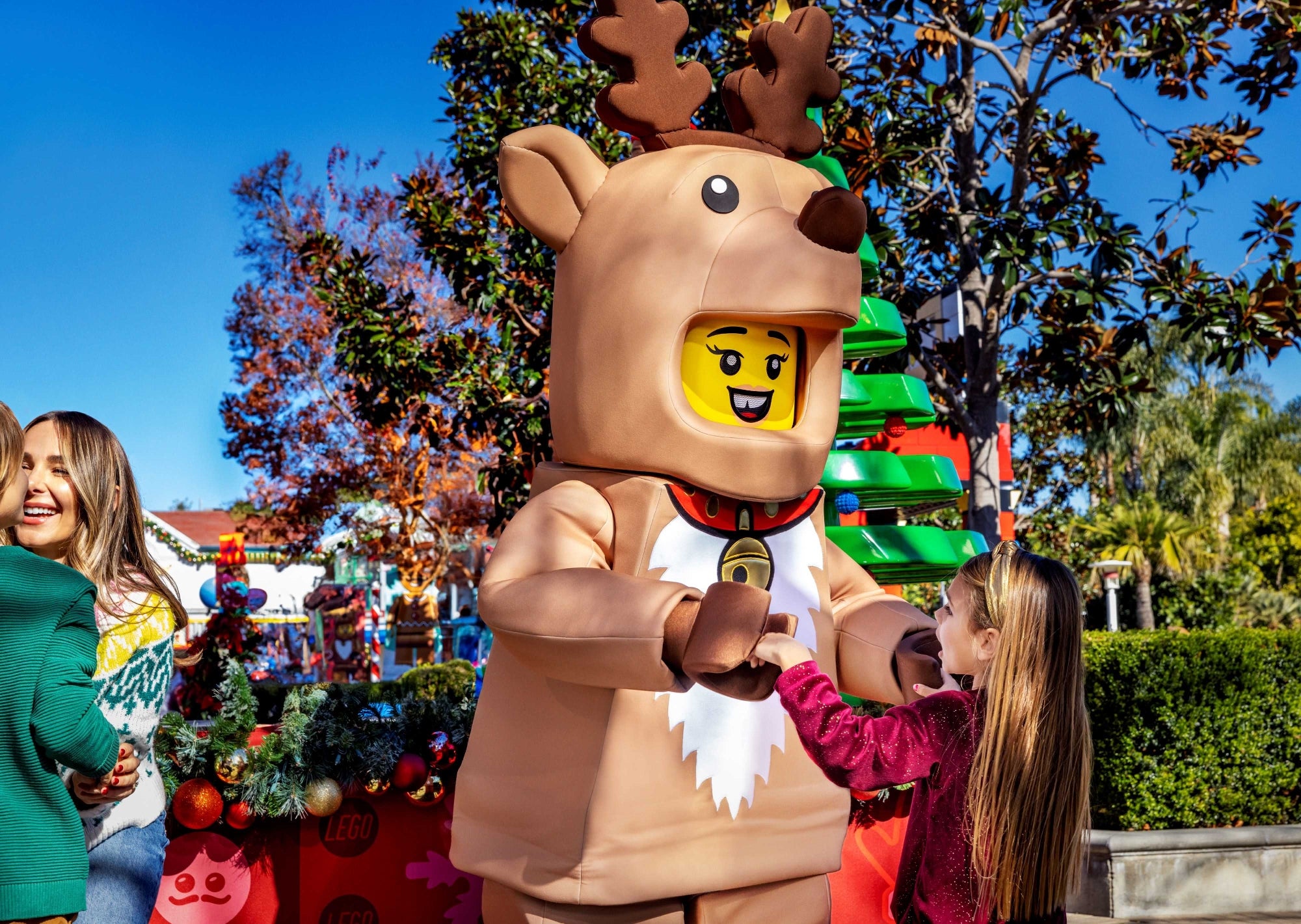 A person in a LEGO reindeer costume interacts with a smiling child outdoors near Christmas decorations and colorful trees.