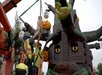 Two workers in safety gear install a large Halloween-themed LEGO sculpture featuring pumpkins, ghosts, and a tree at an outdoor location.