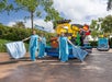 Three people in blue costumes dance with fabric in front of a colorful LEGO parade float featuring a large LEGO figure and ocean-themed decorations, outdoors on a sunny day.
