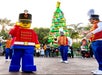Person in a toy soldier costume and musicians in colorful uniforms perform in front of a large, festive LEGO Christmas tree.