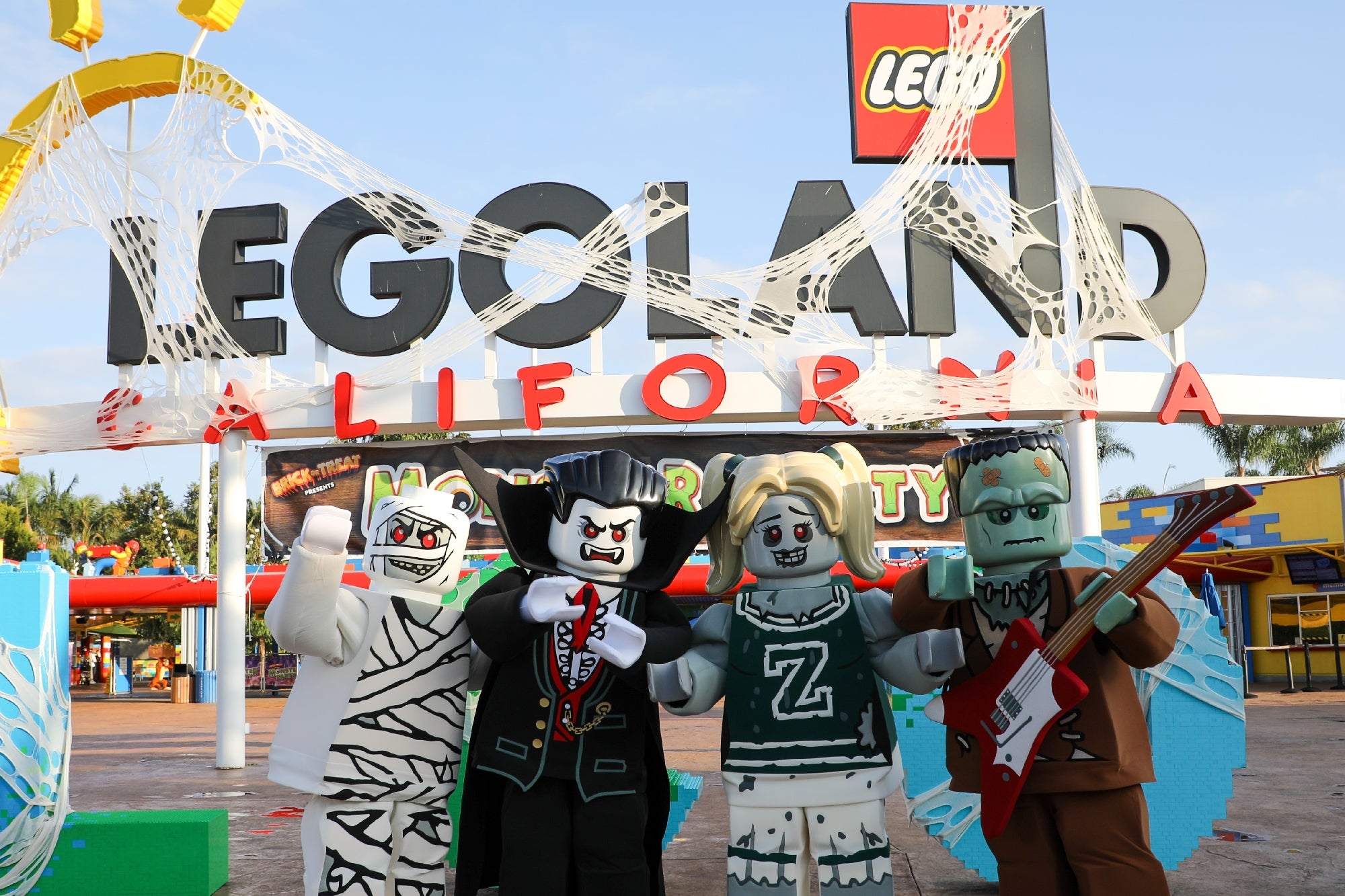 Four people in LEGO monster costumes pose in front of the LEGOLAND California entrance decorated with spider webs.