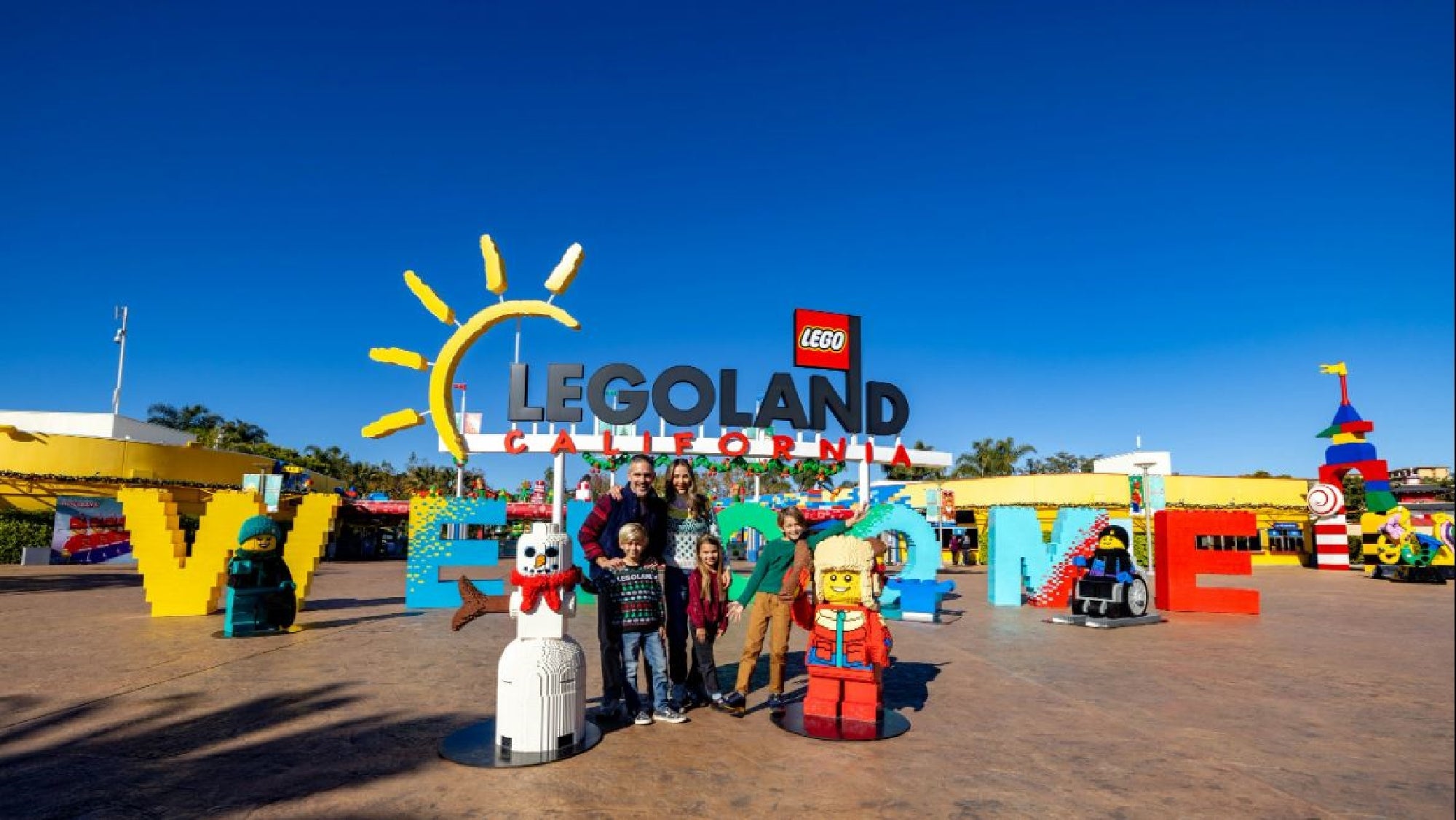 A family poses in front of the Legoland California entrance sign, surrounded by large Lego figures and bright letters spelling "WELCOME.