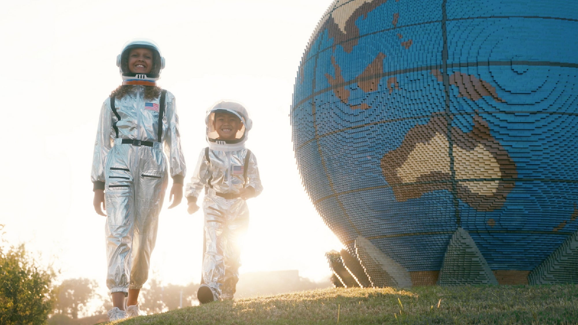 Two children in silver astronaut suits walk outside near a large globe sculpture made of interlocking bricks, with sunlight shining in the background.