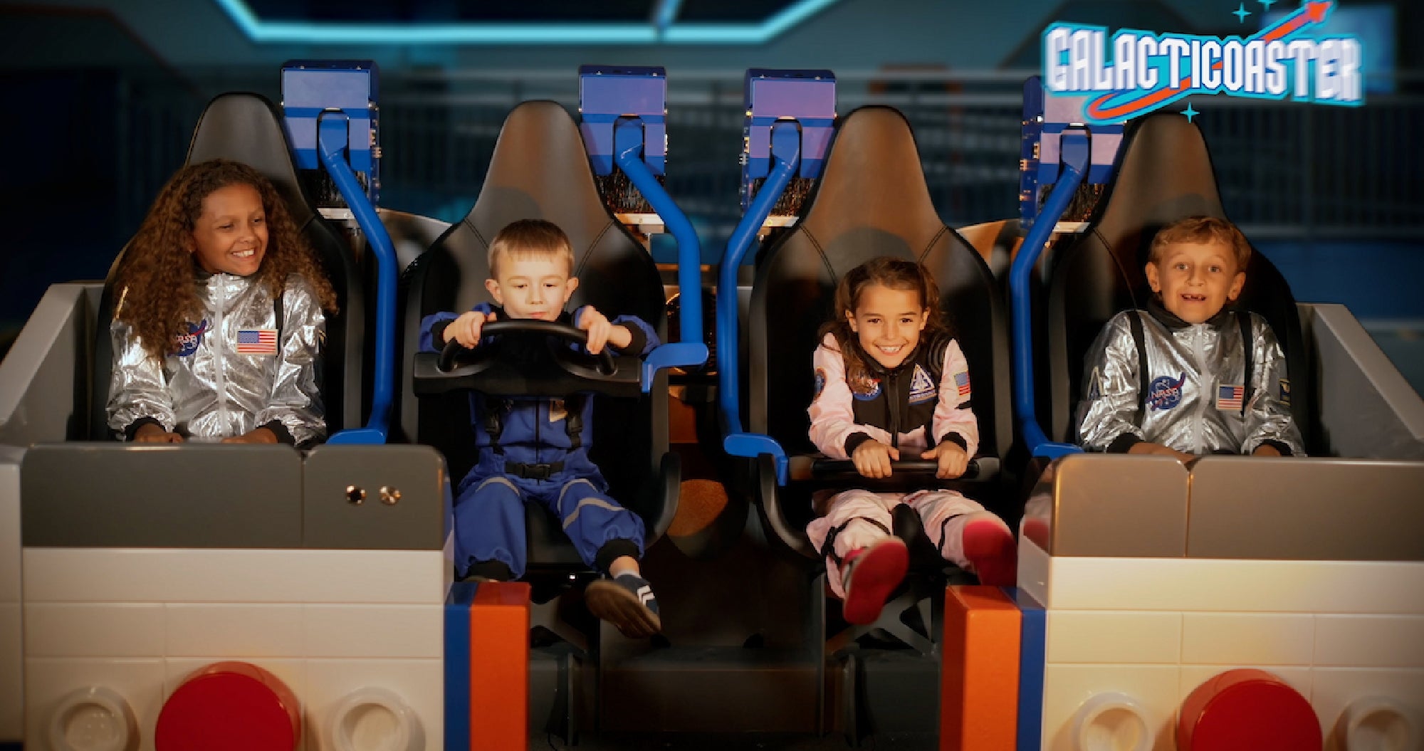 Four children in astronaut outfits sit in a space-themed amusement ride labeled “Galacticoaster,” smiling and ready for the ride to start.