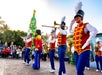 Performers in toy soldier costumes play instruments during an outdoor parade. A large festive tree and crowd are in the background.