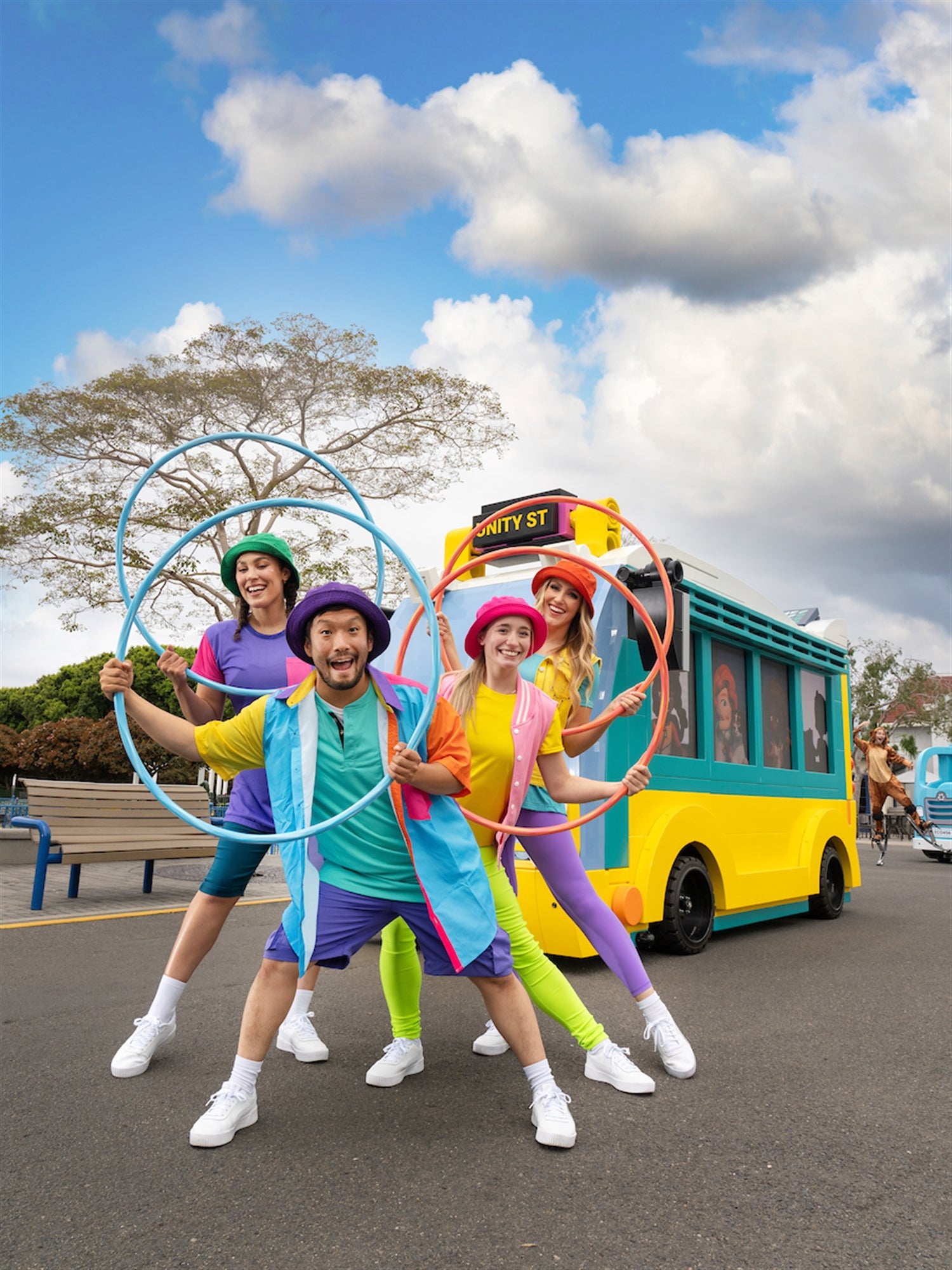Four adults in colorful costumes hold hula hoops and pose in front of a small, bright yellow vehicle on a street during the day.
