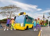 Four performers with colorful outfits and hula hoops stand in front of a whimsical blue and yellow bus marked "Unity St" at a theme park.