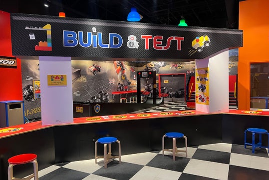 LEGO Build & Test area with a long counter, stools, checkered floor, and colorful decor. The space encourages building and testing LEGO creations.