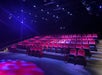 Rows of empty red theater seats face a stage under dim blue and purple lighting in a dark auditorium.
