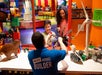 A Master Model Builder demonstrates LEGO models, including a wind turbine and Ferris wheel, to two smiling children at an indoor LEGO exhibit.