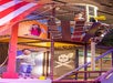 Three children play on a pirate ship-themed indoor playground. One child is on the ground level, while two others are on the upper deck near a yellow telescope. A skull and crossbones flag is visible.