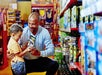 An adult and a child smile while looking at a toy box in a brightly lit toy store aisle filled with colorful boxed toys.