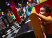 A young child plays with toy cars on a curved ramp in a colorful indoor play area with checkered floors and vibrant decorations.