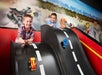 Two boys race toy cars down a black, curved ramp in front of a mural featuring racing cars and a cityscape.