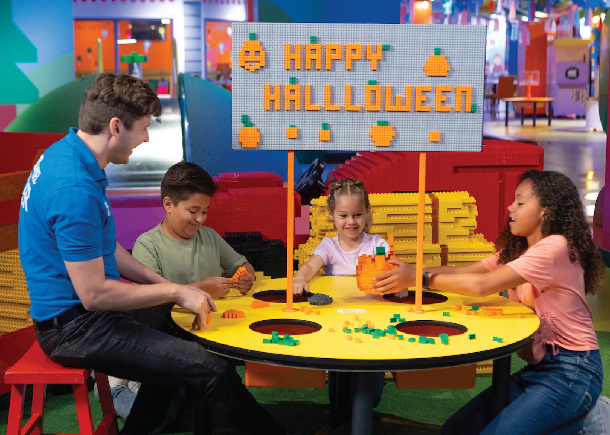 A group of kids and an adult build with toy bricks at a round table under a sign that says "Happy Halloween" with pumpkin decorations.