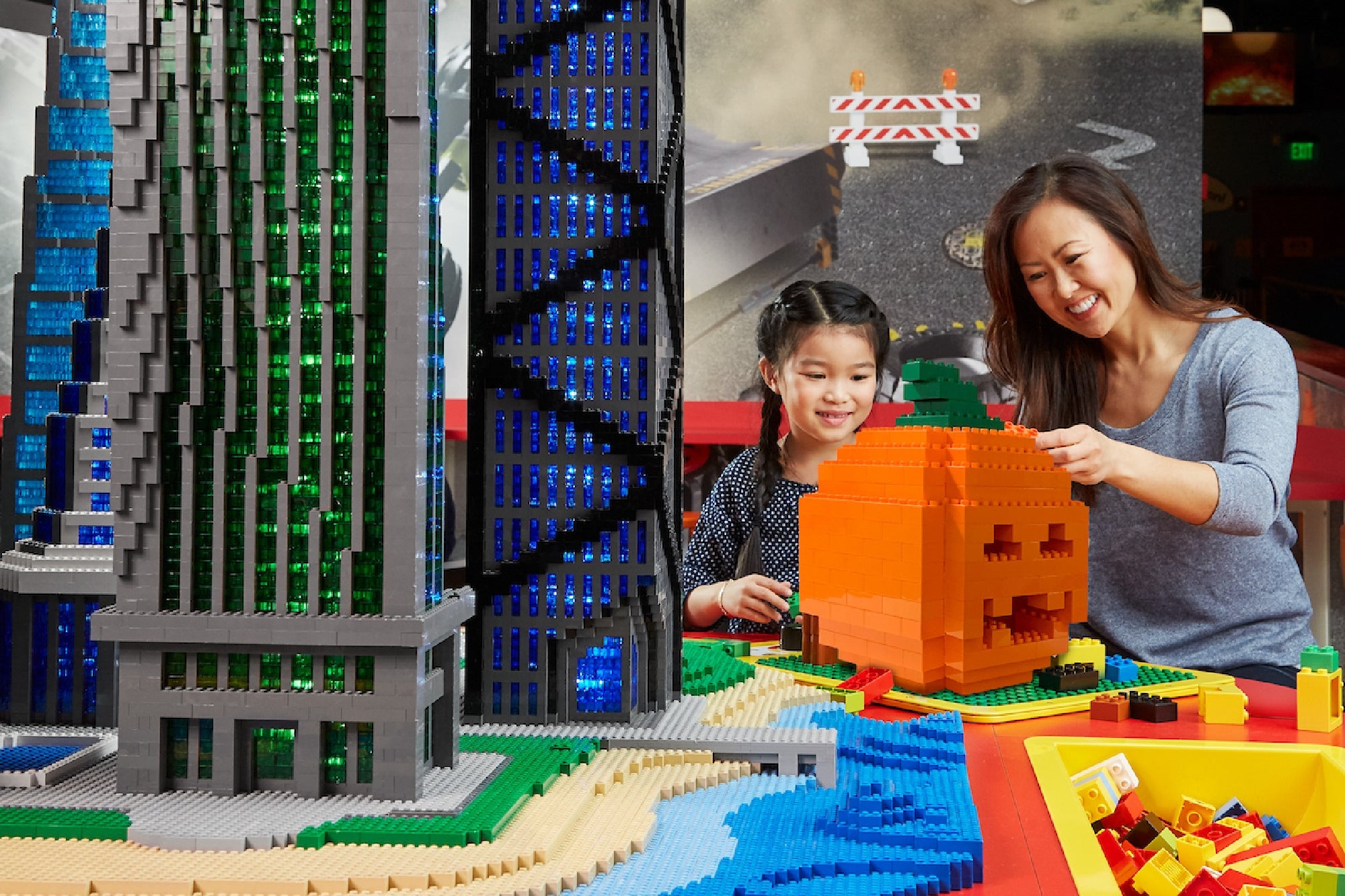 A woman and a young girl build a large orange pumpkin out of LEGO bricks next to a tall LEGO skyscraper model in an indoor play area.