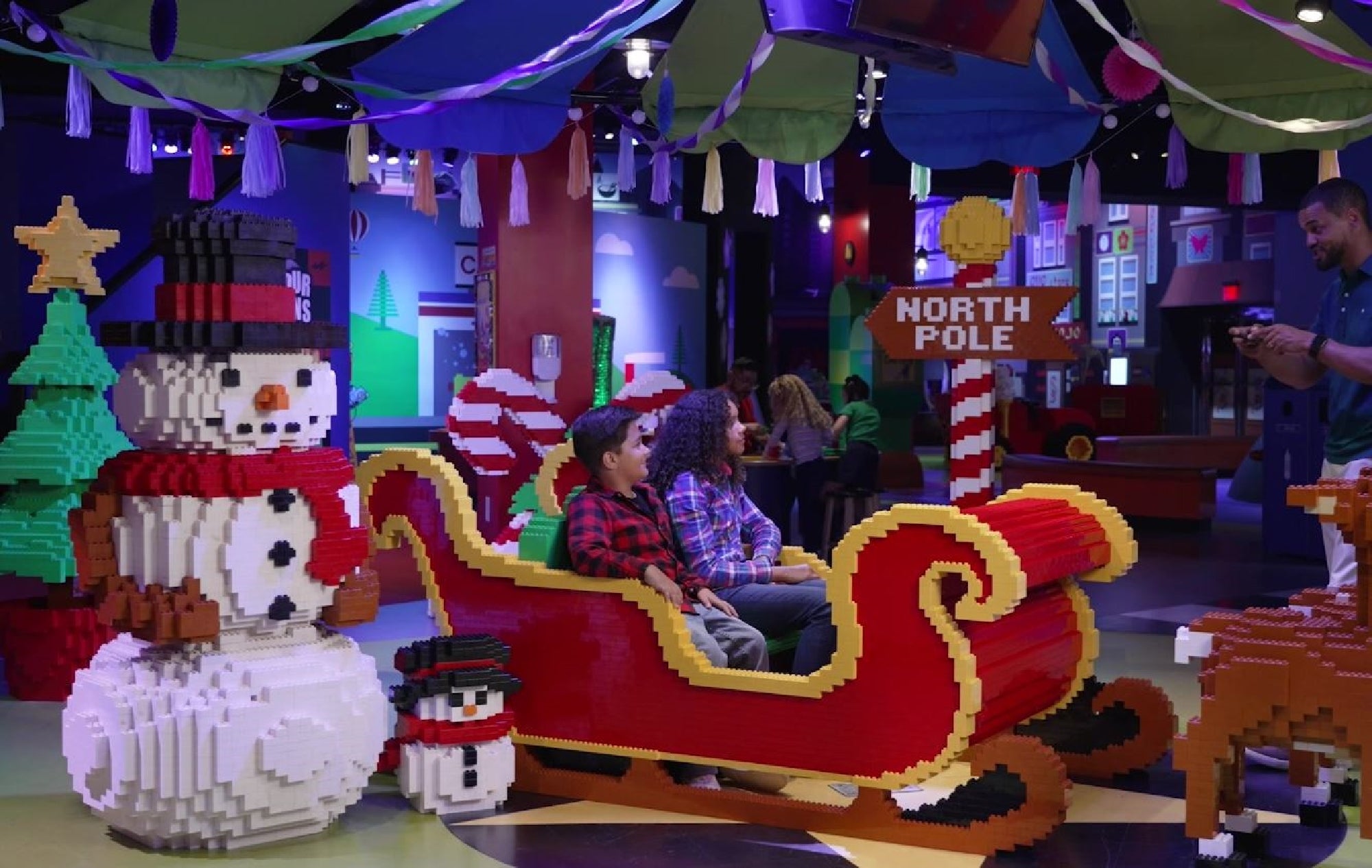 Children sit in a LEGO sleigh display next to a large LEGO snowman and a North Pole sign in a festive, indoor setting while an adult takes their photo.