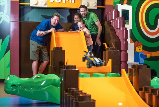 A fun moment at a LEGO-themed play area where two adults and two children joyfully watch a toy car racing down a bright orange slide, with a playful alligator figure in the foreground.