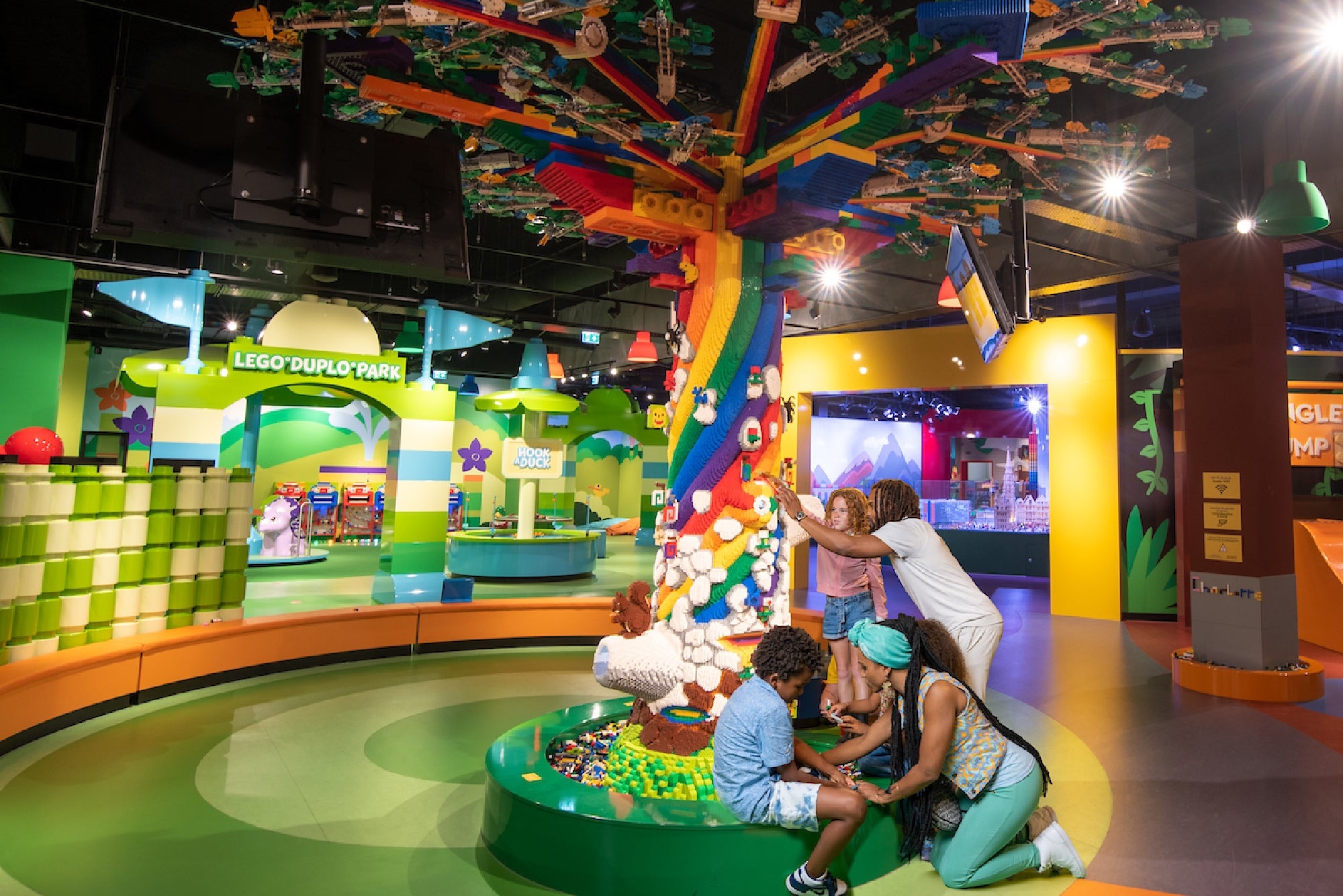 A colorful LEGO-themed play area where children and adults are engaged in building activities around a vibrant tree made of LEGO bricks, with various attractions and decorations in the background.