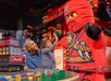 A joyful moment where a young girl is high-fiving a LEGO character dressed as a ninja, while her father watches with a smile, all set against a LEGO-themed backdrop.