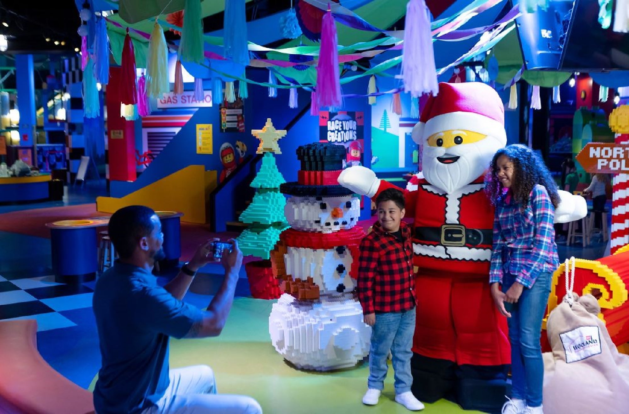 A man takes a photo of two children standing next to a large Santa Claus and snowman made of LEGO bricks inside a colorful indoor play area.