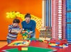 A mother and her son happily building a colorful LEGO structure together, with a playful giraffe made of LEGO bricks visible in the colorful background.