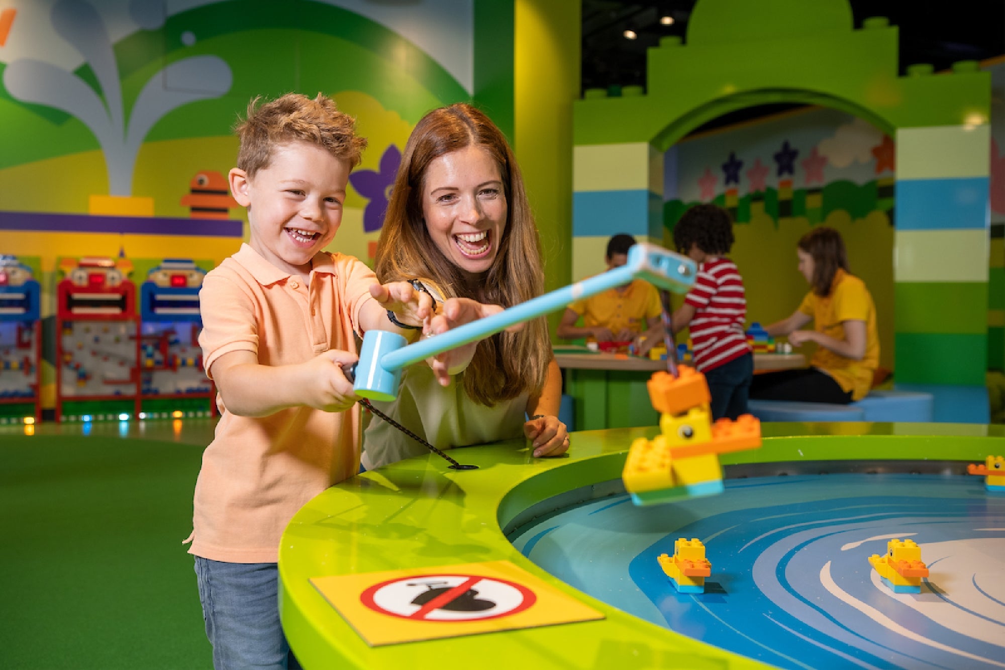A joyful boy and a woman using a fishing rod to catch colorful LEGO ducks in a playful water-themed activity area, surrounded by vibrant decorations and other children engaged in building activities.
