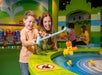 A smiling child and woman use a fishing rod to catch toy ducks at an indoor play area with bright green and yellow decor. Other people play in the background.