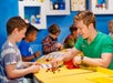 A group of children engaged in a LEGO building activity at a bright, colorful table, with an instructor guiding them as they work on their projects.