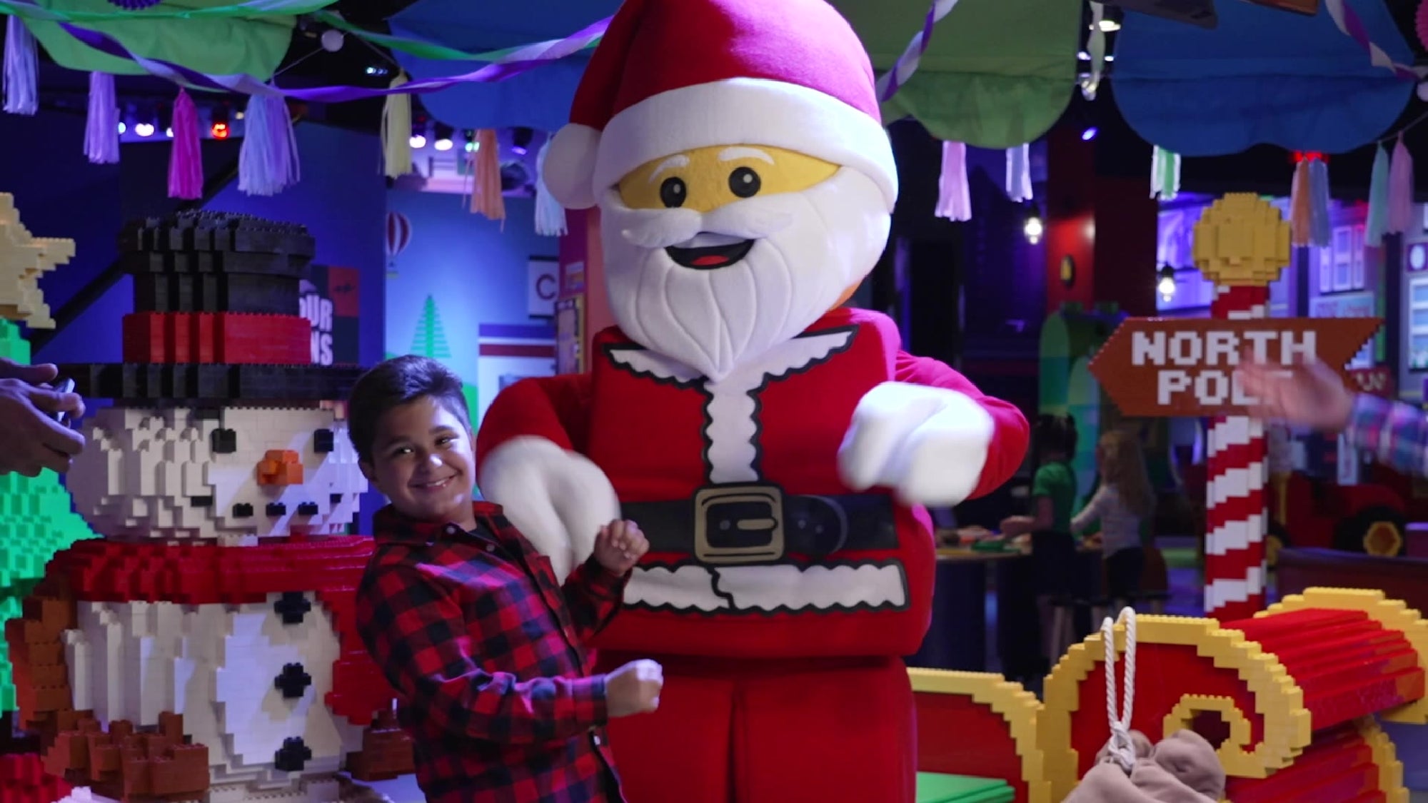 A child poses and smiles next to a person in a large LEGO Santa Claus costume at a festive indoor event with LEGO decorations.