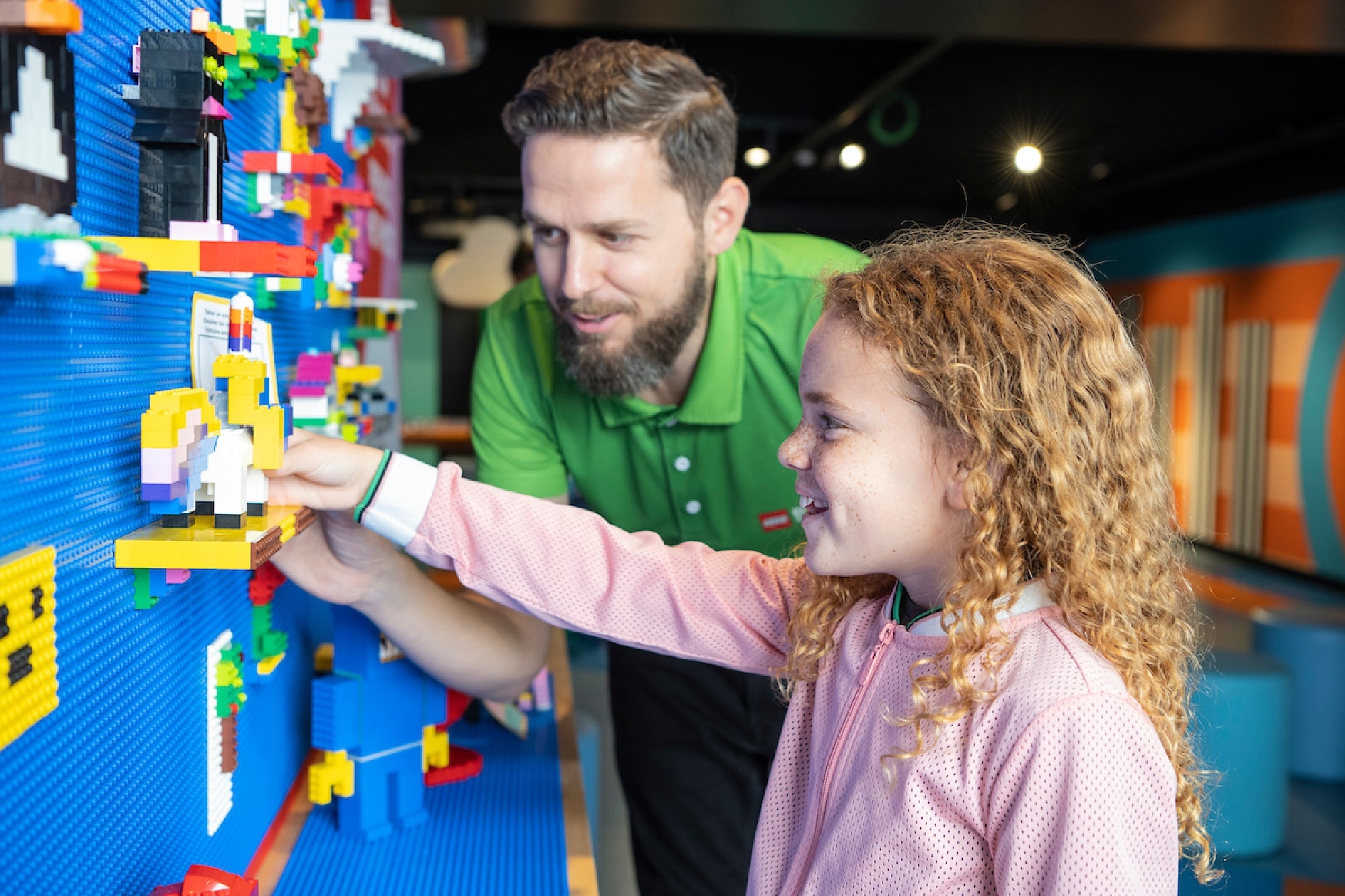 A smiling girl and a man in a green shirt collaborating to add a colorful LEGO figure to a blue wall display filled with various LEGO creations.