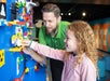 A man and a young girl build with colorful LEGO bricks on a vertical blue wall in a brightly lit play area.