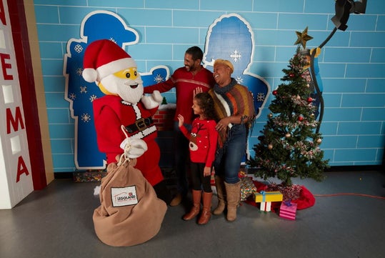 A family poses and smiles with a person in a LEGO Santa costume next to a decorated Christmas tree and a bag labeled "LEGOLAND.